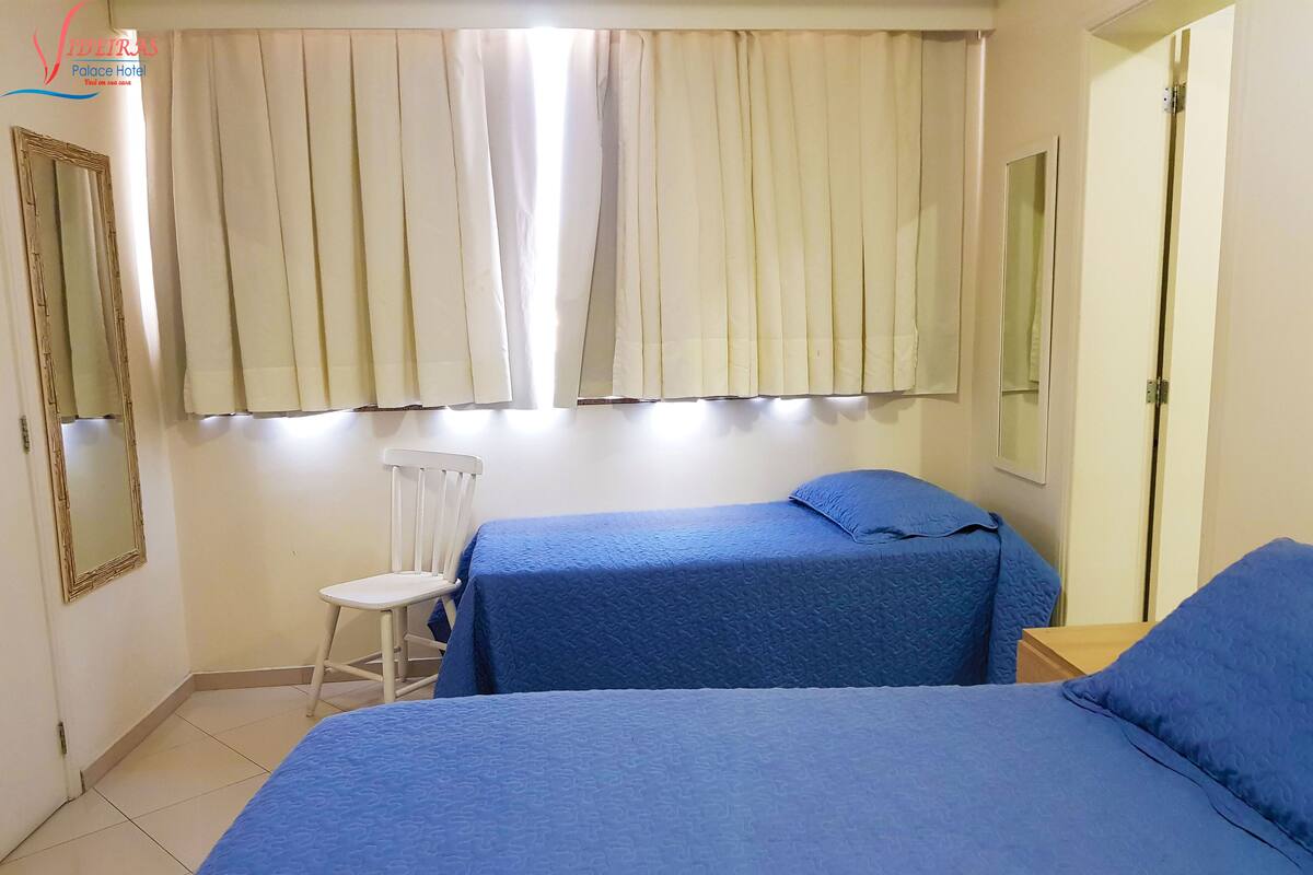 Een kamer met twee bedden, een stoel, een spiegel en een raam met gordijnen.