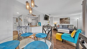 A modern living room with a sofa, ottoman, and coffee table, a kitchen with white cabinets, and a dining area with chairs and a glass table.