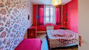 A room with floral wallpaper, a bed with a patterned bedspread, a red dresser, and a window with curtains.