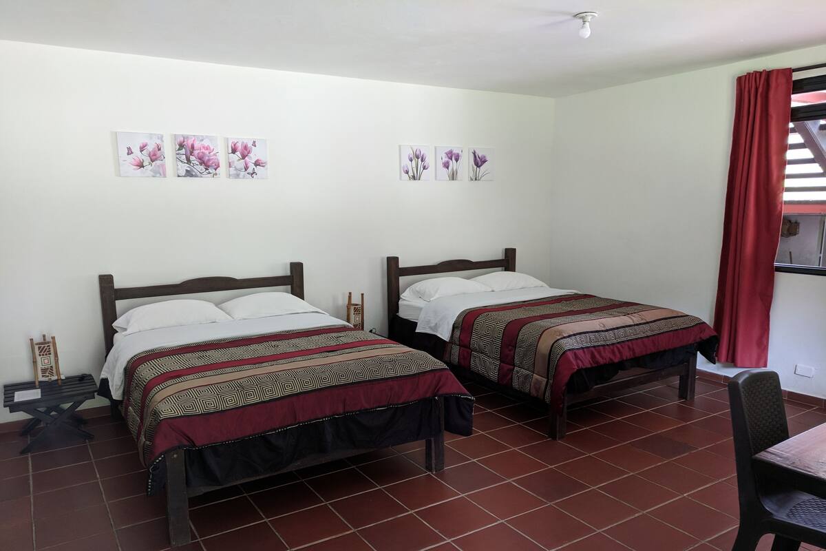 A room with two beds, each with patterned bedding, a small table, and a chair.