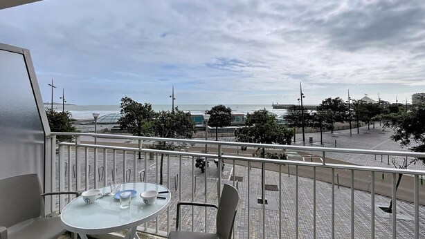 Un balcon avec une table et des chaises, donnant sur un espace pavé agrémenté d’arbres et offrant une vue sur la mer.