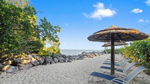 A beach with lounge chairs and thatched umbrellas, a rocky shoreline, and a clear blue sky. -  Mariner's Club 606 • Clean & Reliable 4/4 • Amazing Pools & Resort Amenities (Key Largo)