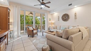 A spacious living room with a sofa, a coffee table, and a dining area. There is a ceiling fan and a large window with curtains. -  Mariner's Club 606 • Clean & Reliable 4/4 • Amazing Pools & Resort Amenities (Key Largo)
