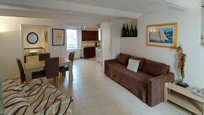 Een moderne woonkamer met een bruine bank, een eethoek met een tafel en stoelen, een keuken op de achtergrond, en een foto in een lijst aan de muur.