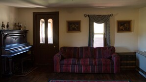 A living room with a dark wooden piano, a patterned sofa, and two framed pictures on the wall. - Ranch House in the Elkhorn Mountains (Clancy)