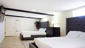 A bedroom with two beds, a dark headboard, a bedside table with a lamp, and a door leading to another room.
