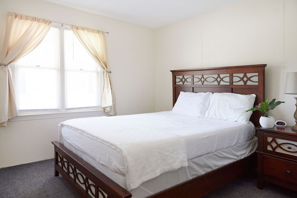 A neatly made bed with a wooden headboard, a bedside table with a clock, and a window with curtains.