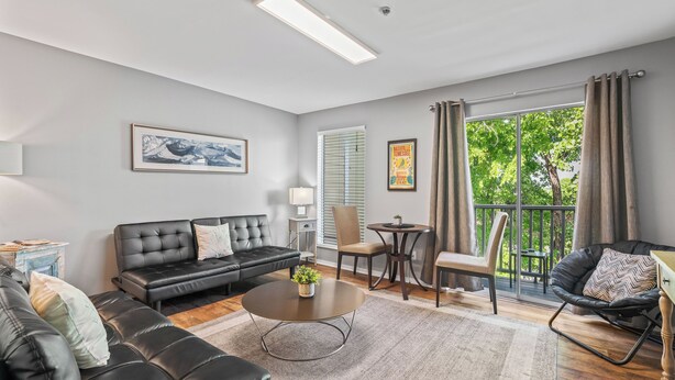 A modern living room with a black leather sofa, a round coffee table, and a black leather chair. There is a small table with a lamp and a chair, and a large window with curtains. - Captain's Quarters Riverfront #1 (Nashville)
