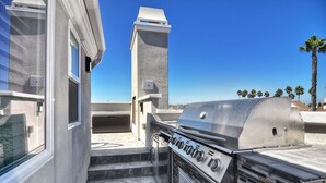 An outdoor kitchen area with a stainless steel grill, a stone fireplace, and a view of palm trees. - Newly built + Ocean View + Steps to Sand (Newport Beach)