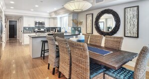 A modern kitchen with white cabinets, a central island, and a dining area with wicker chairs and a wooden table. - Newly built + Ocean View + Steps to Sand (Newport Beach)