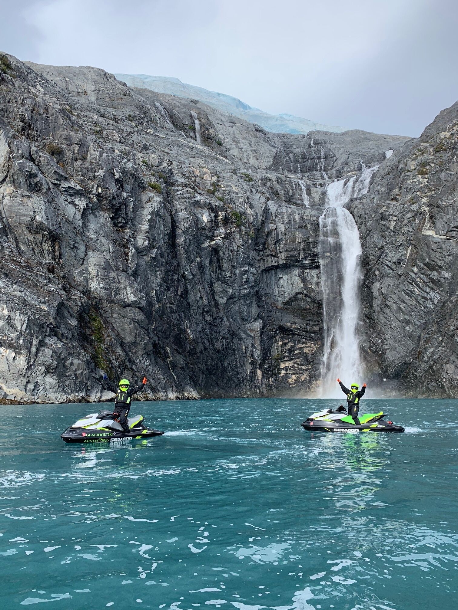 Twee personen op jetski's in het water, met een waterval op de achtergrond.