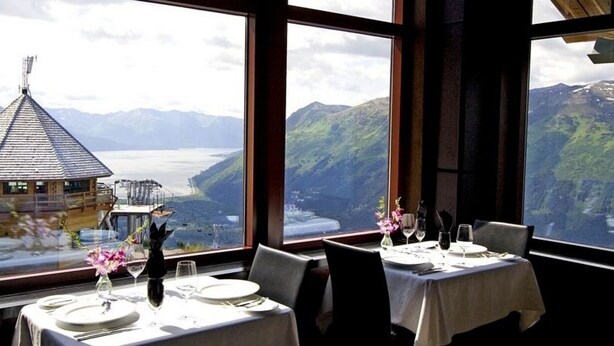 Un espace repas avec des tables préparées pour deux personnes, offrant une vue sur un paysage montagneux avec un lac et une structure en bois en arrière-plan.