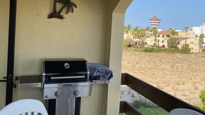 A balcony with a grill, a palm tree decoration, and a view of a beach and buildings.