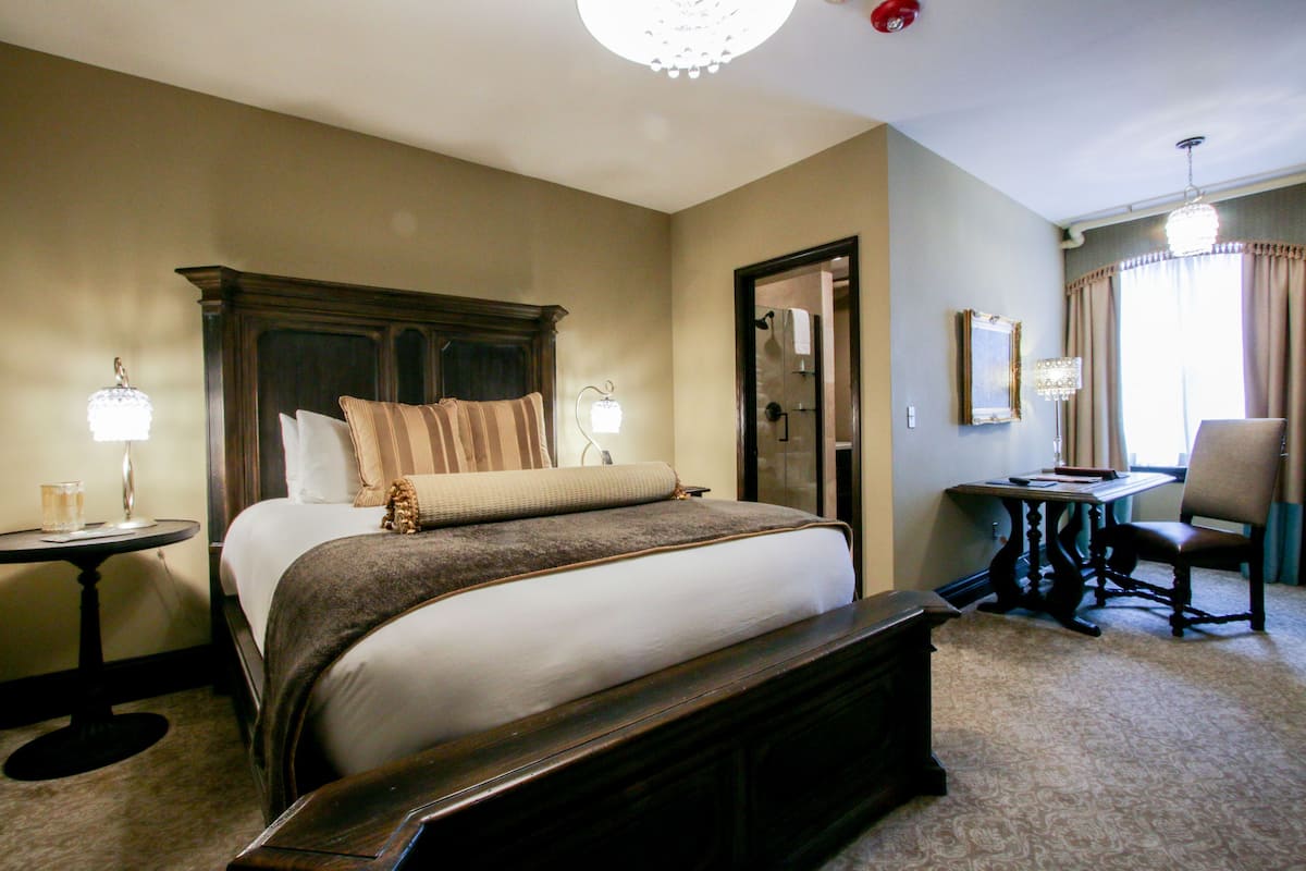 A hotel room with a large bed, a bedside table with a lamp, a chair, a small table, and a mirror on the wall.