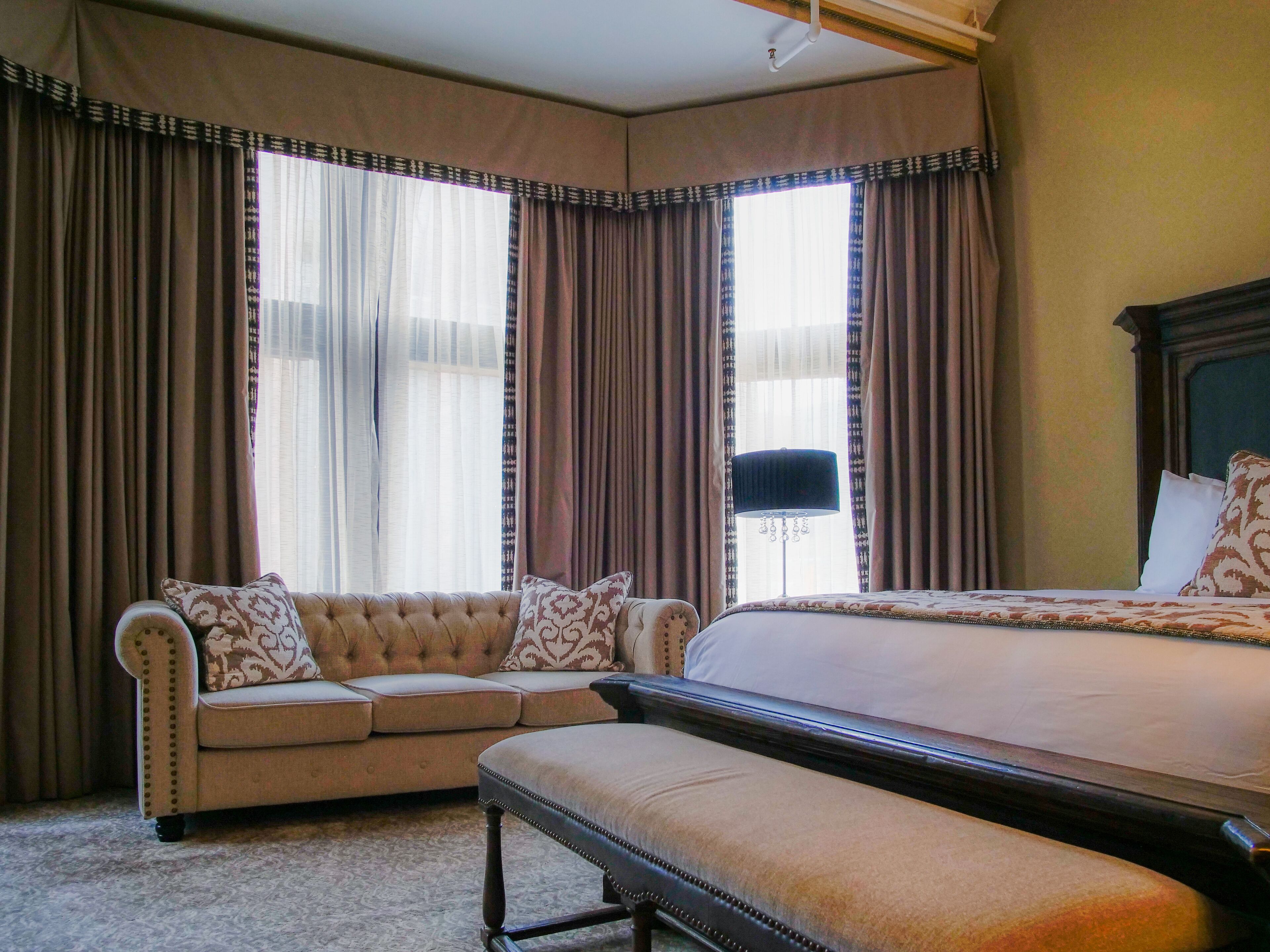 Un dormitorio con una cama grande, un sofá y un banco. Hay cortinas en las ventanas y se ve una lámpara.