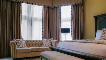 Un dormitorio con una cama grande, un sofá y un banco. Hay cortinas en las ventanas y se ve una lámpara.