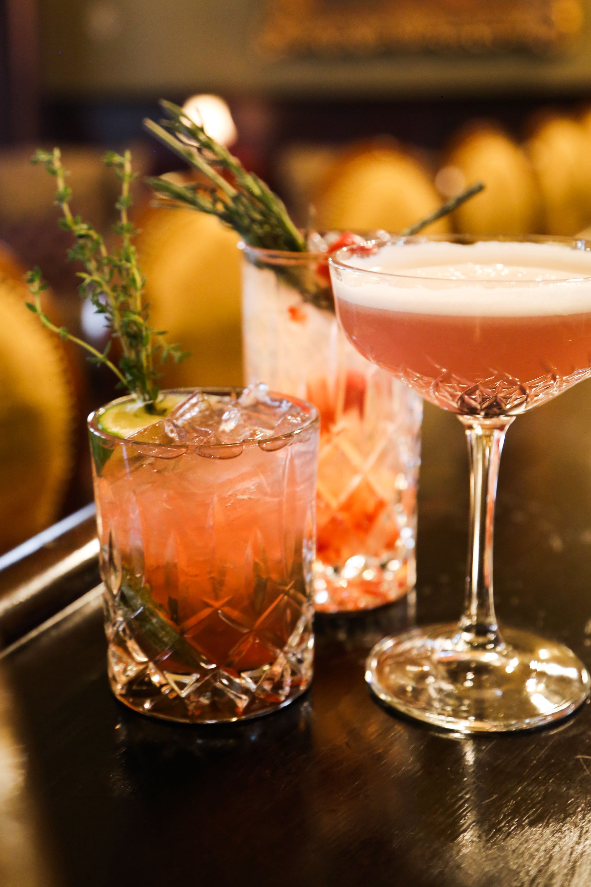 Two cocktails in glasses with ice cubes and garnishes on a wooden bar counter.