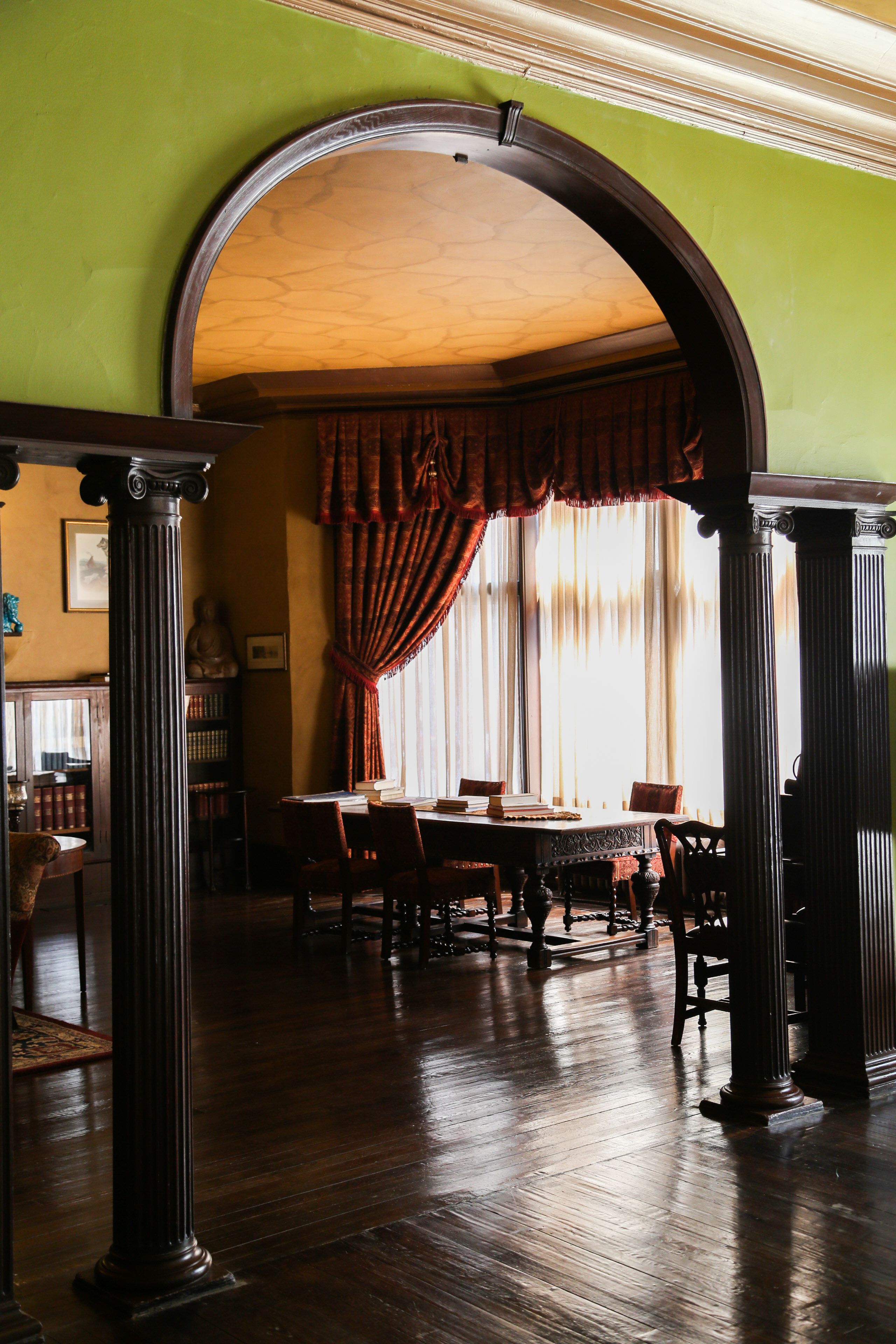 A dining area with a wooden table and chairs, a large window with curtains, and a green wall.