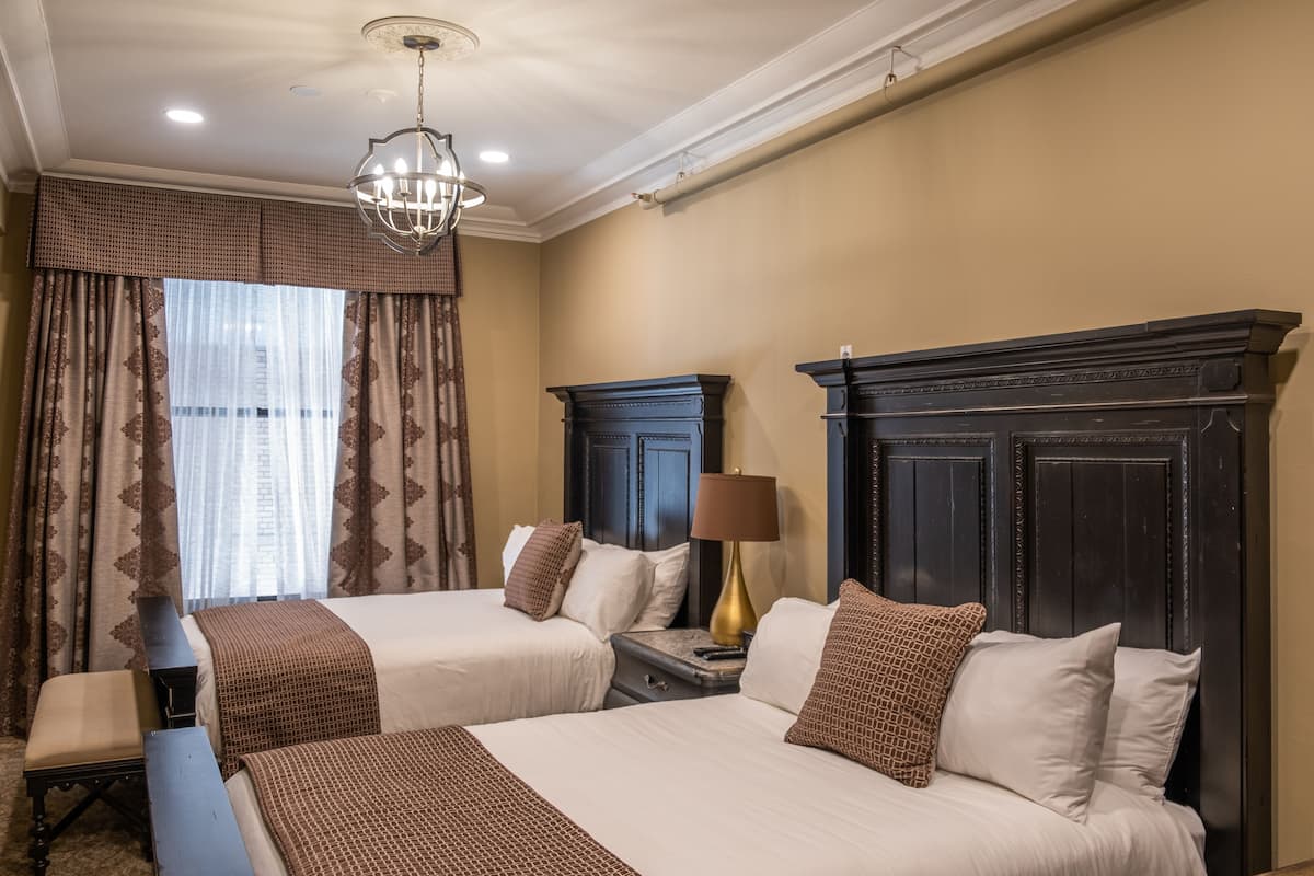 A hotel room with two beds, a chandelier, a fireplace, and a window with curtains.