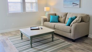 Un salon avec un canapé beige, une table basse et un tapis à rayures. Il y a une fenêtre avec des stores, une lampe sur une table d’appoint et un tableau au mur.