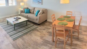 Un salon comprenant un canapé, une table basse et une table à manger avec des chaises. On y trouve également un tableau au mur, un ventilateur de plafond et un appareil de climatisation.