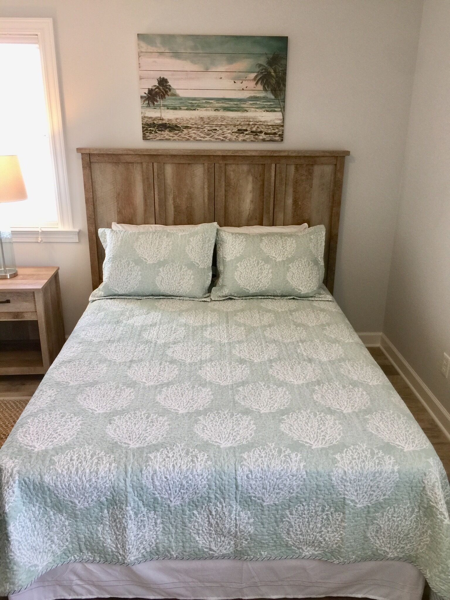 Una cama con una colcha celeste estampada y dos almohadas a juego, un cabecero de madera, una mesita de noche con una lámpara y un cuadro con una escena de playa sobre la cama.