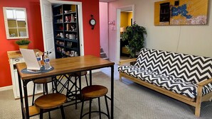 Un coin repas comprenant une table en bois, deux tabourets, une bibliothèque et un canapé agrémenté d’un coussin à motifs chevrons.