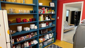 Une cuisine avec des étagères bleues remplies de divers ustensiles et appareils de cuisine, un chat noir et blanc sur une des étagères, et un mur rouge en arrière-plan.