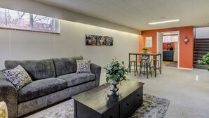 Un salon avec un canapé gris, une table basse noire et une plante. On y trouve également un coin repas avec une table et des chaises, et une cuisine à l’arrière-plan.