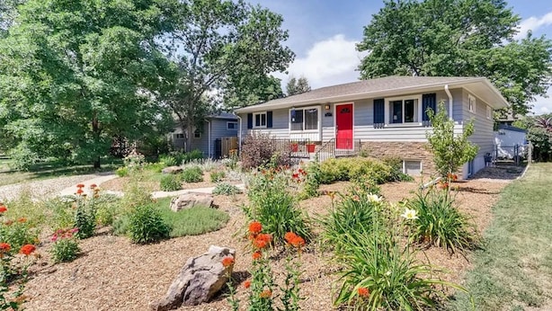 Une maison de plain-pied avec une porte rouge, entourée d’un jardin bien entretenu où poussent différentes plantes et fleurs.