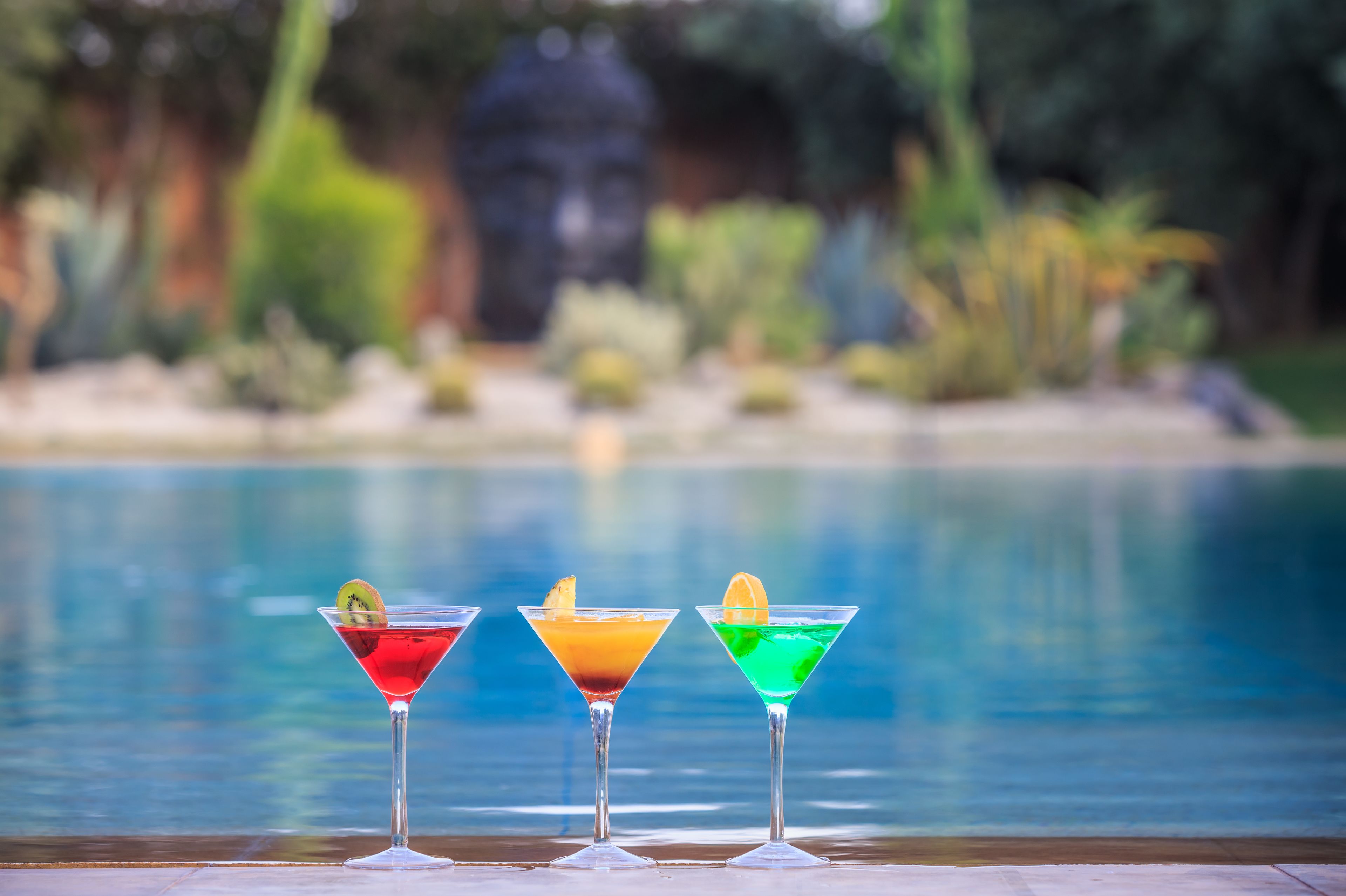 Three martini glasses with colorful drinks and garnishes by a poolside.