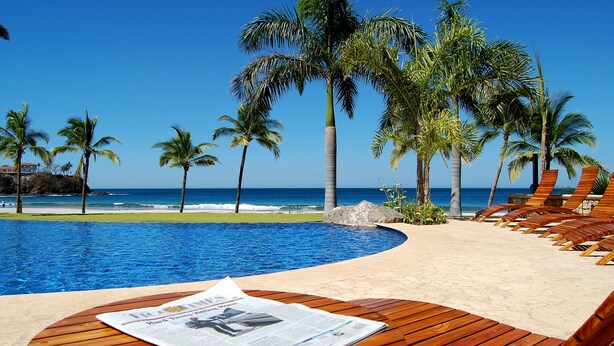 A poolside area with wooden loungers, a newspaper, and a view of the ocean and palm trees.