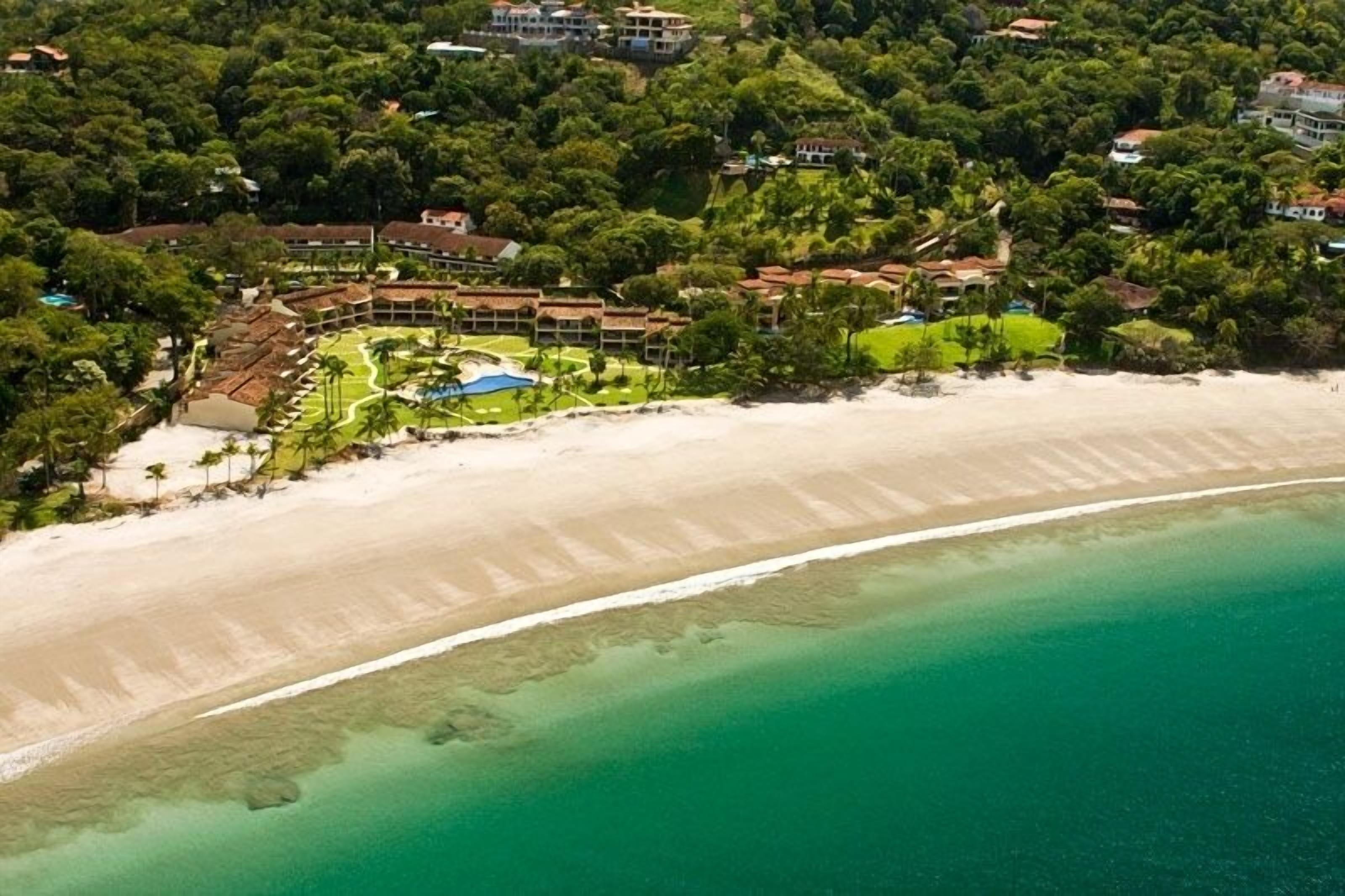 Vue aérienne d’une station balnéaire avec une plage de sable, des eaux turquoise et limpides, et une végétation luxuriante.