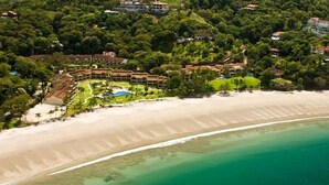 An aerial view of a coastal resort with a sandy beach, clear turquoise waters, and lush greenery.