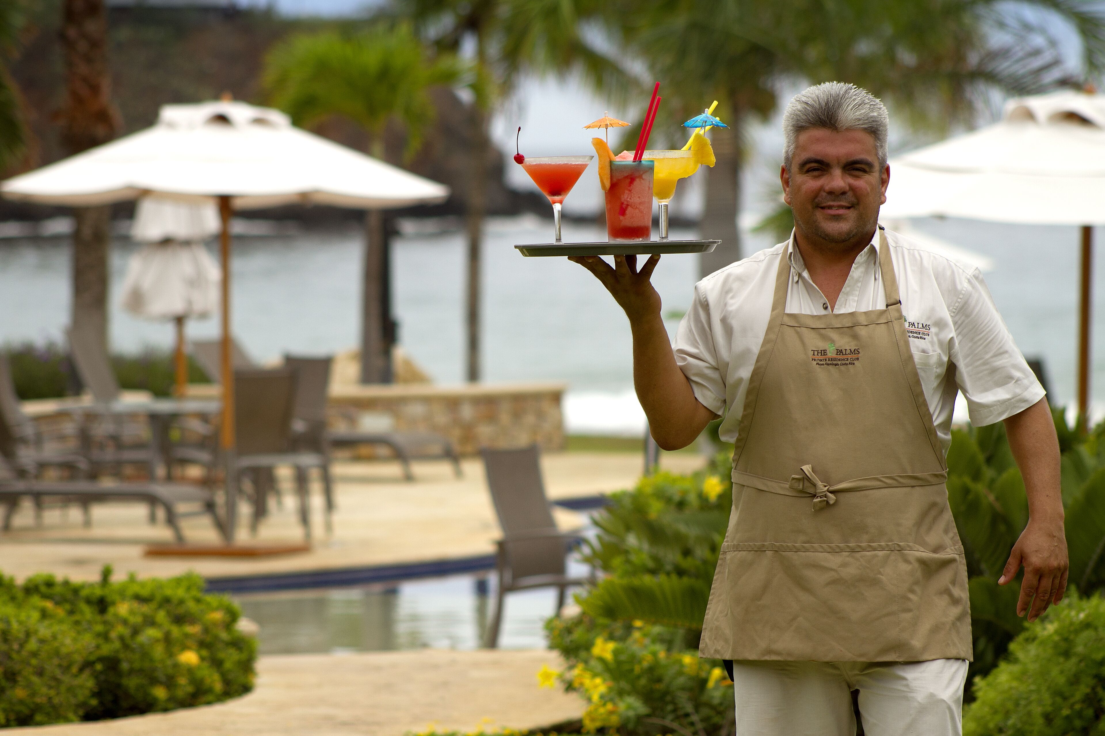 A person holding a tray with two cocktails and a fruit garnish, standing by a pool with lounge chairs and umbrellas.