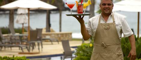 A person holding a tray with two cocktails and a fruit garnish, standing by a pool with lounge chairs and umbrellas.