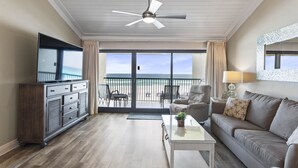 Een ruime woonkamer met een plafondventilator, een flatscreen-tv, een bank, een salontafel en een balkon met uitzicht op het strand.