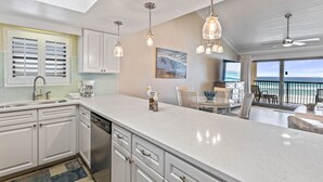 Een moderne keuken met witte kastjes, een groot eiland, hangende lampen en uitzicht op de oceaan via het balkon.
