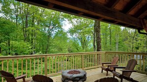 A wooden deck with Adirondack chairs and a stone fire pit, overlooking a forested area. - Creekfront Cabin | Hot Tub | 1GB WiFi | Gated | Pets Welcome (Maggie Valley)