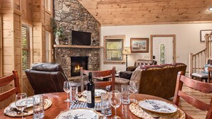 A cozy living room with a stone fireplace, leather furniture, and a dining table set for six. - Creekfront Cabin | Hot Tub | 1GB WiFi | Gated | Pets Welcome (Maggie Valley)