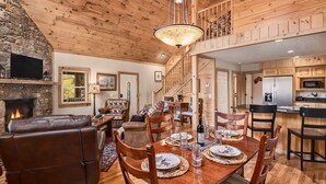 A spacious living room with a fireplace, wooden ceiling, and a dining area with a wooden table and chairs. - Creekfront Cabin | Hot Tub | 1GB WiFi | Gated | Pets Welcome (Maggie Valley)