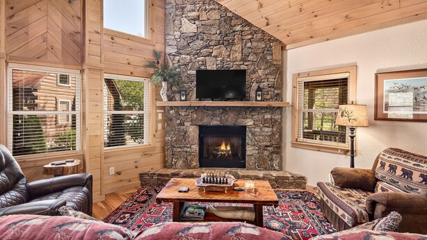 A cozy log cabin interior with a stone fireplace, wooden walls, and a large window overlooking a forest. - Creekfront Cabin | Hot Tub | 1GB WiFi | Gated | Pets Welcome (Maggie Valley)