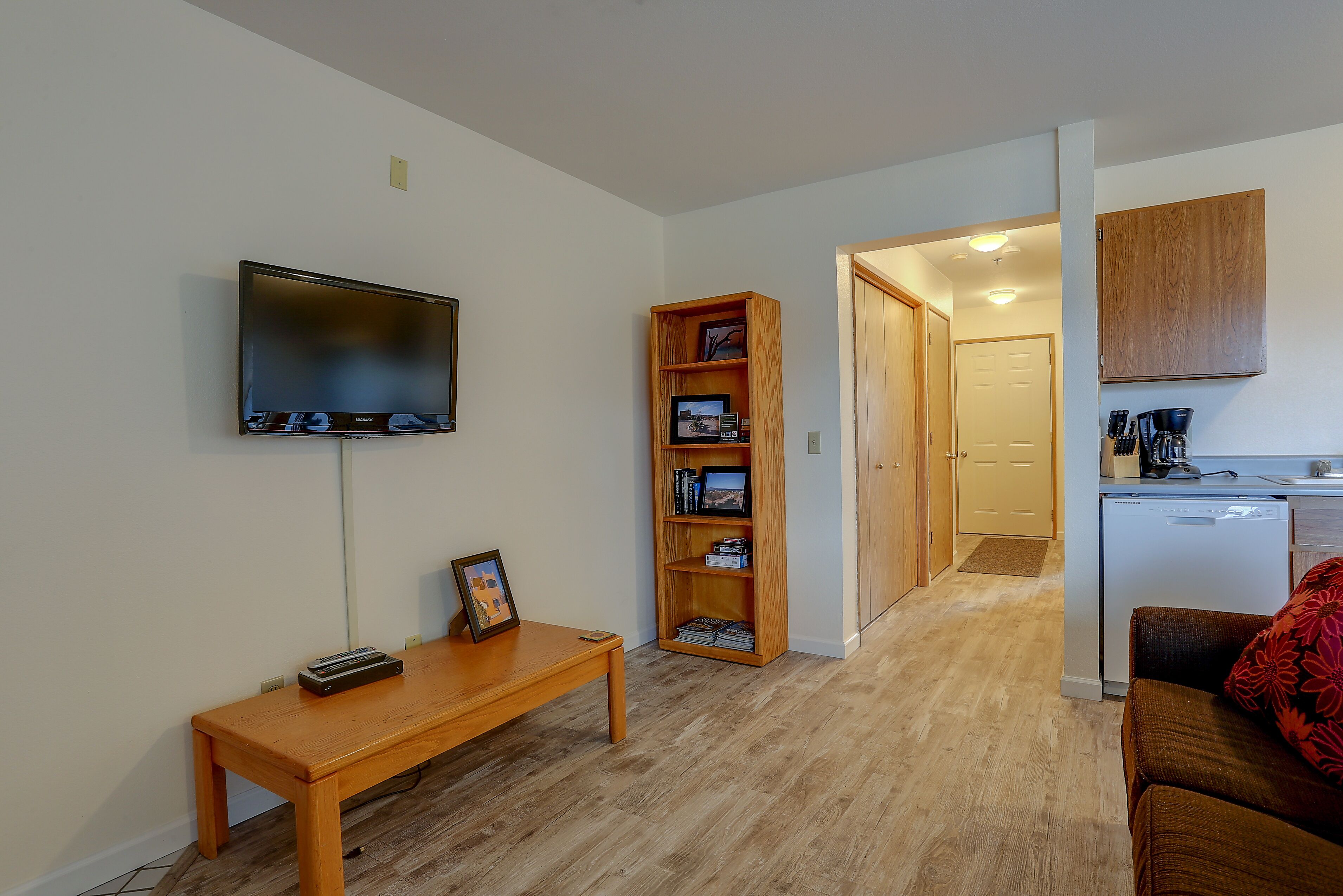 Una sala de estar con un televisor de pantalla plana, una estantería de madera, un banco de madera y una cocina con un refrigerador y armarios.