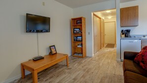 Een woonkamer met een flatscreen televisie, een houten boekenkast, een houten bankje en een keuken met een koelkast en kasten.