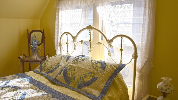 A bedroom with a bed, a window with lace curtains, a chair, and a decorative vase.