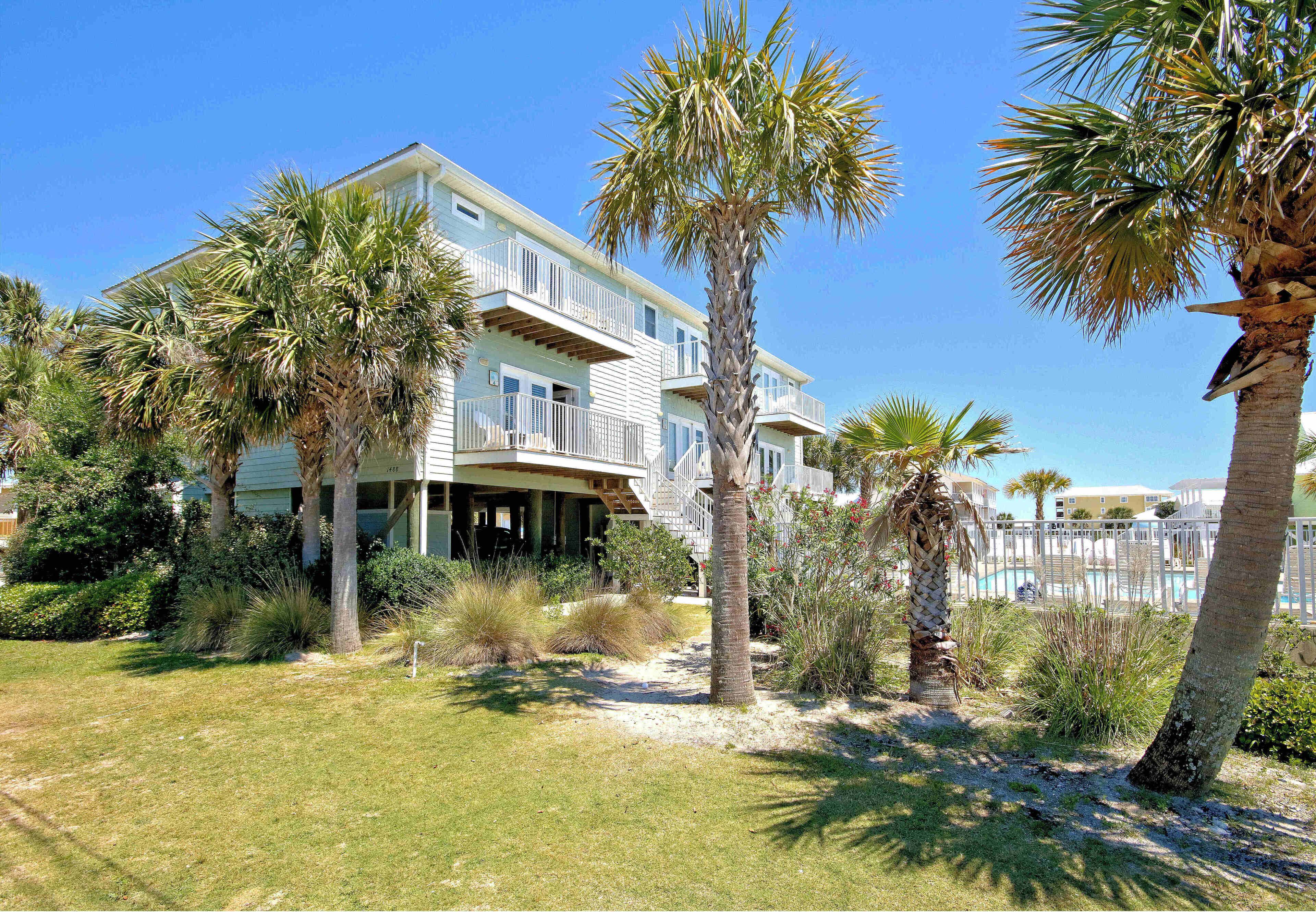A two-story beachfront house with a balcony, surrounded by palm trees and a sandy lawn.
