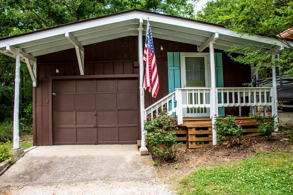 A single-story building with a brown garage door, a porch with a white railing, and a small garden area with pink flowers. - On the Rocks Cottage. Lakefront, Dog-Friendly. Sleeps 16. Bunkhouse. Free Wifi (Eagle Rock)