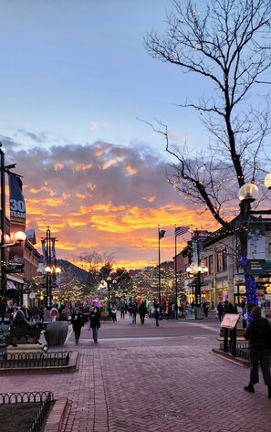 A public square with a sunset in the background, featuring a statue, brick pavement, and illuminated street lamps.