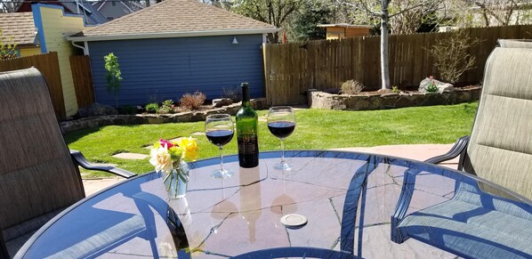 A round glass table with two wine glasses and a bottle, surrounded by outdoor furniture.