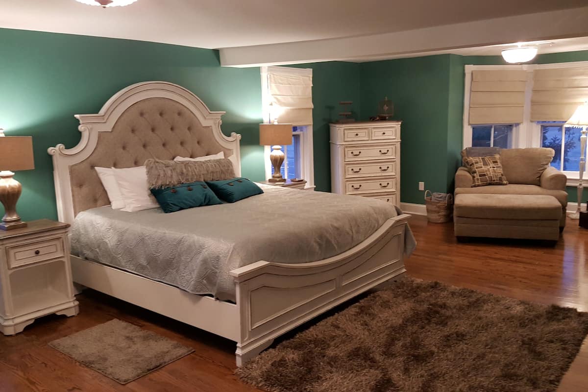 A bedroom with a large bed, a nightstand, a dresser, a chair, and a window with blinds.